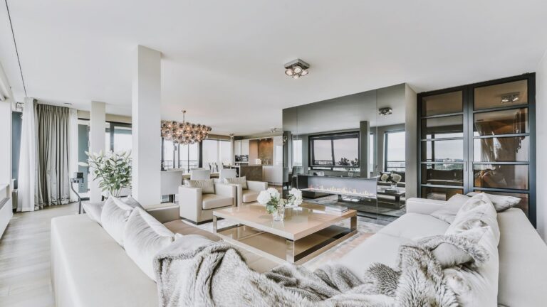 Open-plan living room with white sofas, a glass coffee table, and a linear fireplace, plus large windows and a mirrored wall.