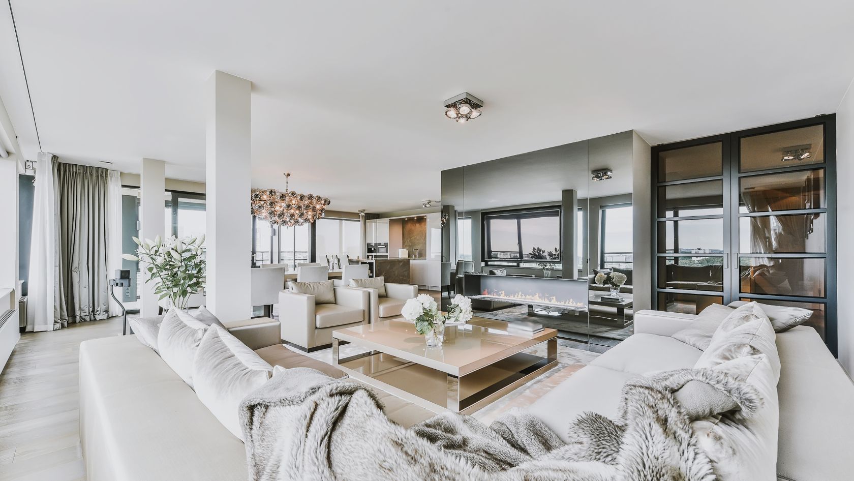 Open-plan living room with white sofas, a glass coffee table, and a linear fireplace, plus large windows and a mirrored wall.