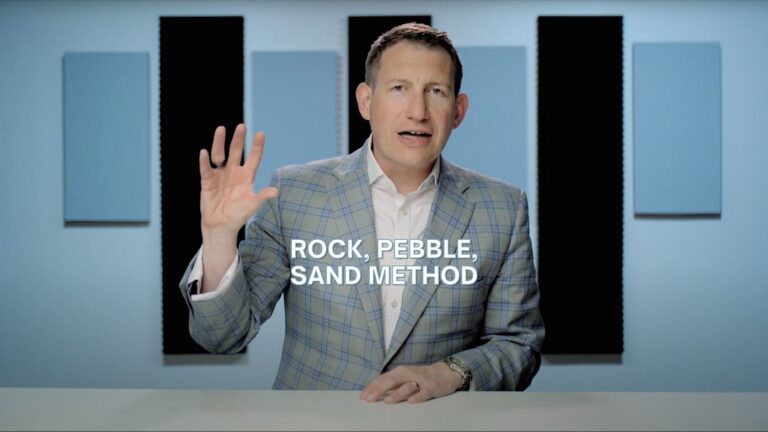 Presenter in a light plaid blazer speaks to the camera in a studio with blue acoustic panels; caption reads 'ROCK, PEBBLE, SAND METHOD'.