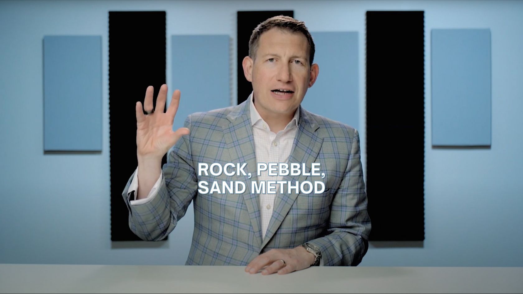 Presenter in a light plaid blazer speaks to the camera in a studio with blue acoustic panels; caption reads 'ROCK, PEBBLE, SAND METHOD'.