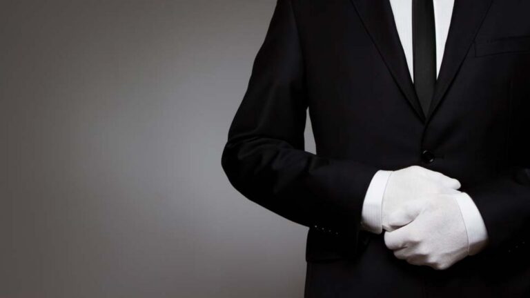 Person wearing a black suit and white gloves, hands folded in front.