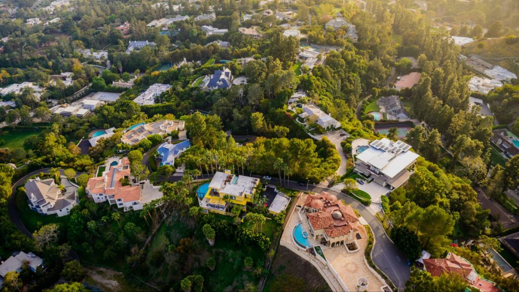 Aerial view of a hillside suburb with large houses, winding roads and many trees with pool-filled backyards nearby.