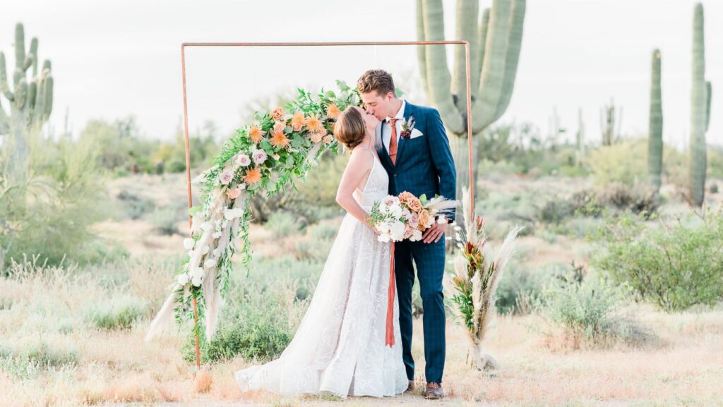Bride and groom kiss beneath a copper-framed floral arch in a desert with tall cacti in the background, holding a bouquet of peach and pink flowers.