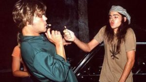 Two young men outside at night; one with a beanie lights a cigarette for the other beside a car.