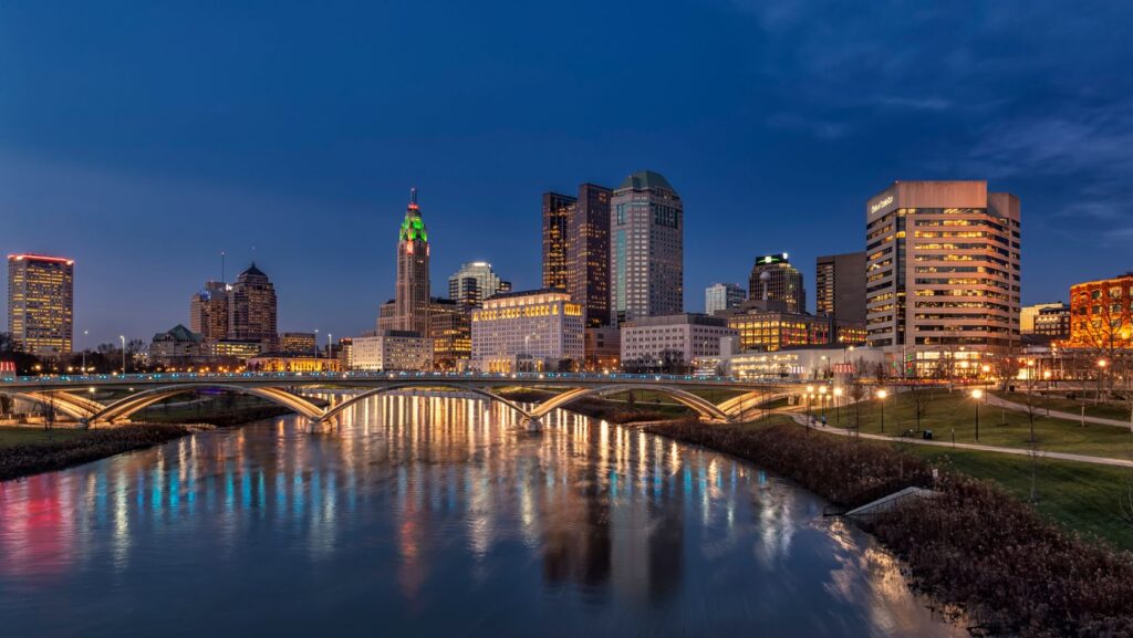 City skyline at dusk with illuminated skyscrapers, a lit bridge, and reflections on a calm river.