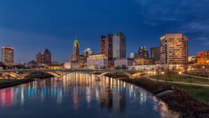 City skyline at dusk with illuminated skyscrapers, a lit bridge, and reflections on a calm river.