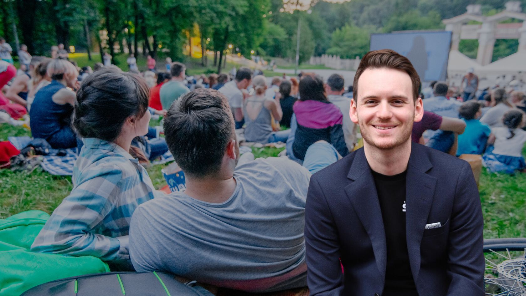 Smiling man in a dark blazer sits at the edge of a park crowd during an outdoor event.