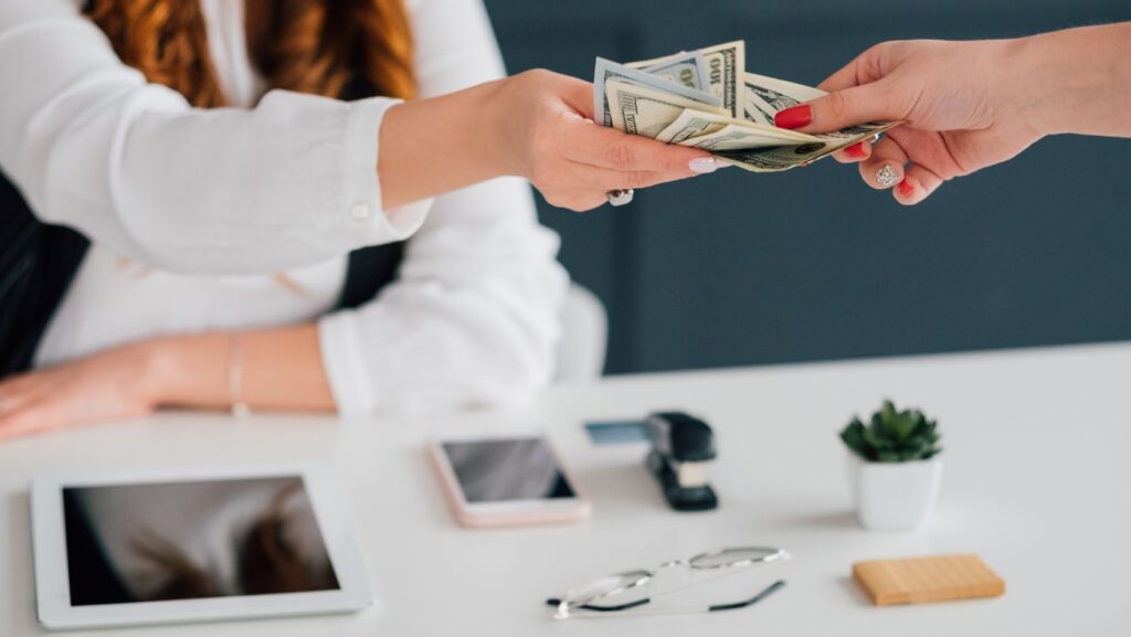 Two hands exchanging a stack of dollar bills across a white desk, suggesting a payment or cash transaction in progress.