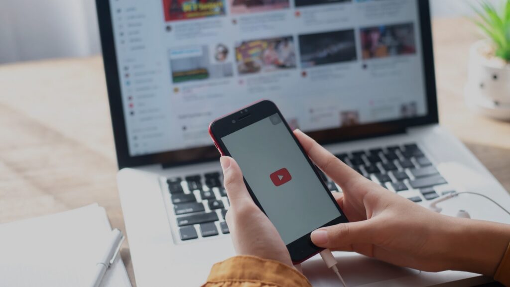 Person holding a smartphone with the YouTube app open, while a laptop screen shows YouTube videos/feed in the background.