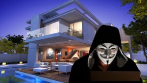 Hooded figure wearing a white Guy Fawkes mask sits with a laptop beside a modern glass-walled house at dusk by a pool area.