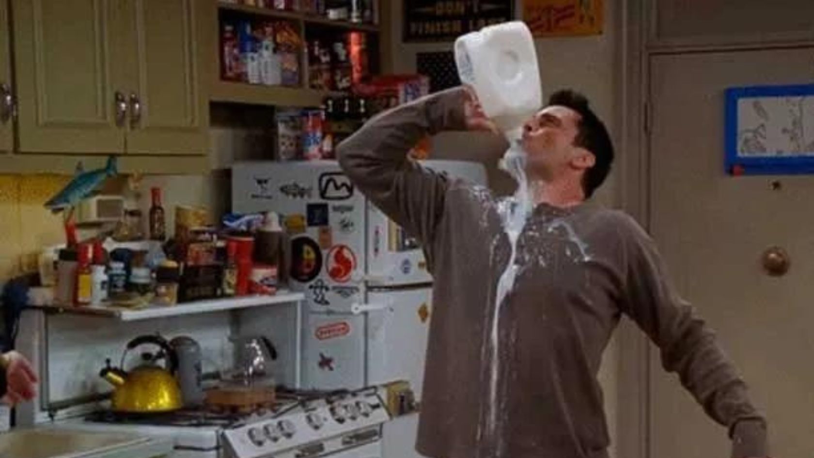Man in a brown shirt tilts a large milk jug over his mouth, milk spilling down his shirt in a cluttered kitchen.