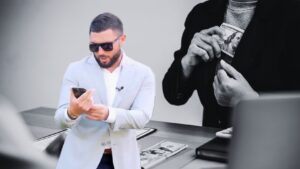 Man in a light blazer using a smartphone in the foreground; a grayscale image of hands holding money appears in the background, suggesting wealth.
