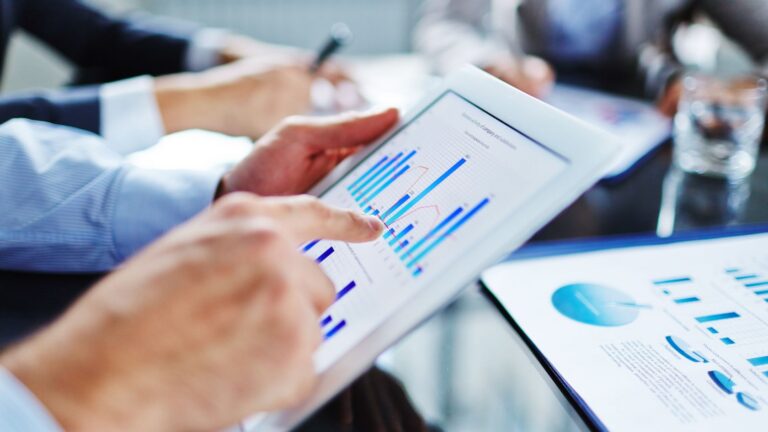 Hands of professionals pointing at a tablet displaying blue bar charts during a meeting.
