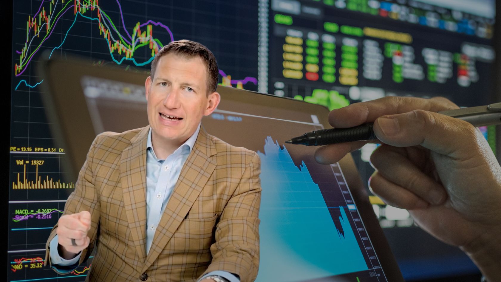 Businessman in a beige plaid blazer explains stock charts on a tablet as a hand with a stylus points at a blue graph in front of market screens.