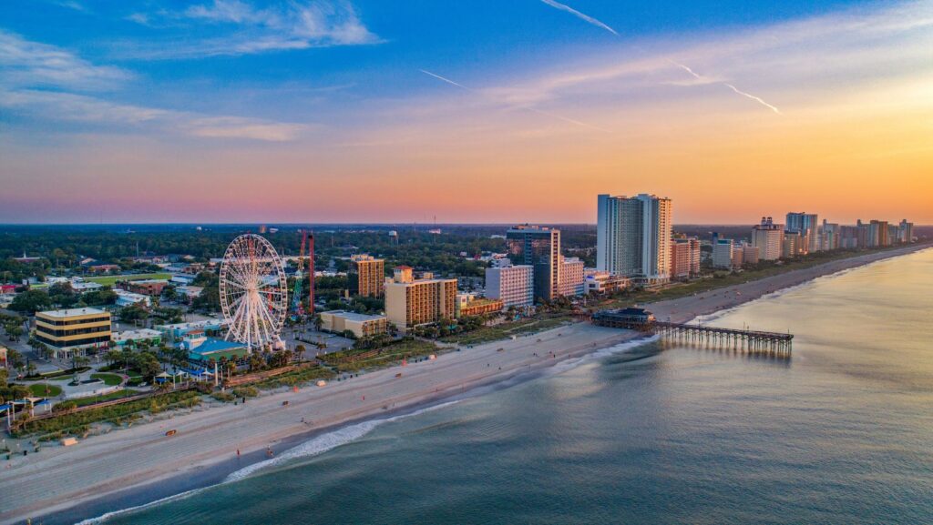 Aerial view of a sunny coastal city with a ferris wheel, high‑rise hotels, and a long beachside pier along the water.