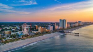Aerial view of a sunny coastal city with a ferris wheel, high‑rise hotels, and a long beachside pier along the water.