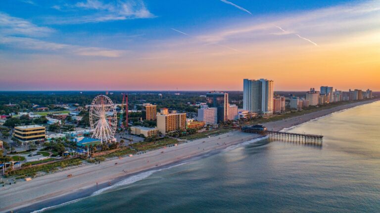 Aerial view of a sunny coastal city with a ferris wheel, high‑rise hotels, and a long beachside pier along the water.