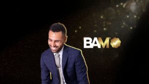 Smiling man in a blue plaid suit outlined in gold, beside bold 'BAM' with a gold clover and check mark on a starry black background.