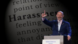 Speaker at a podium giving a talk, with a large printed page showing the word 'Harassment' in the background