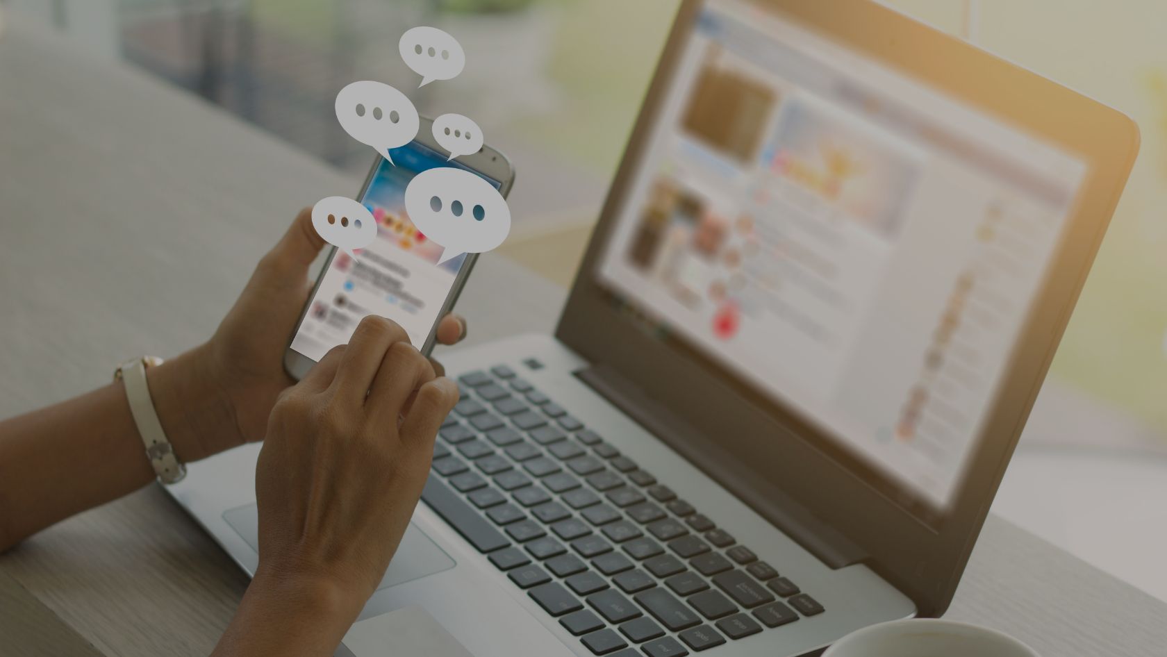 Person typing on a smartphone with chat bubbles floating above, in front of an open laptop show­ing a webpage.