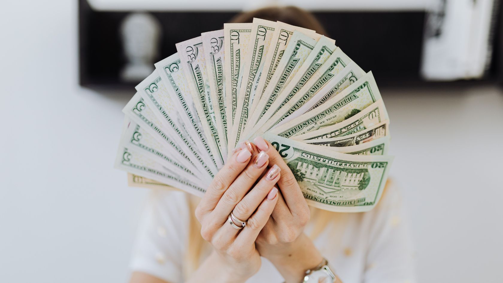 Person hiding their face behind a fan of US dollar bills held in both hands, showing pink nails and rings in the foreground on a blurred background