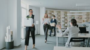 Three professionals walk through a bright office carrying moving boxes; the woman in front waves toward a seated colleague.