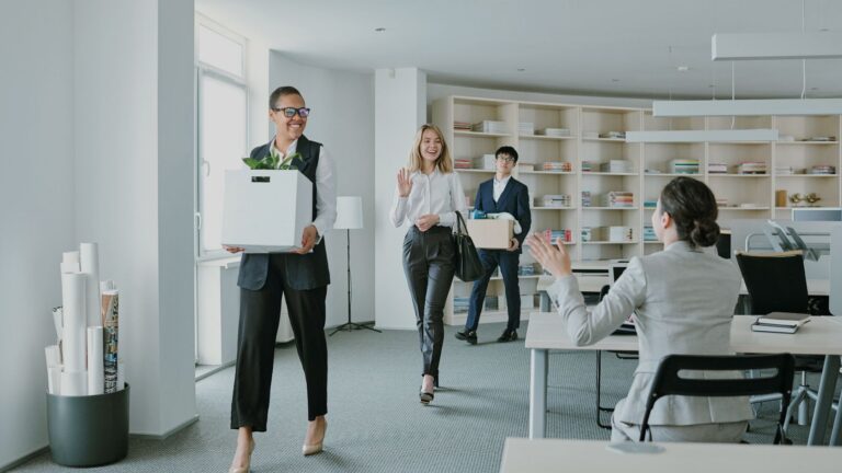 Three professionals walk through a bright office carrying moving boxes; the woman in front waves toward a seated colleague.