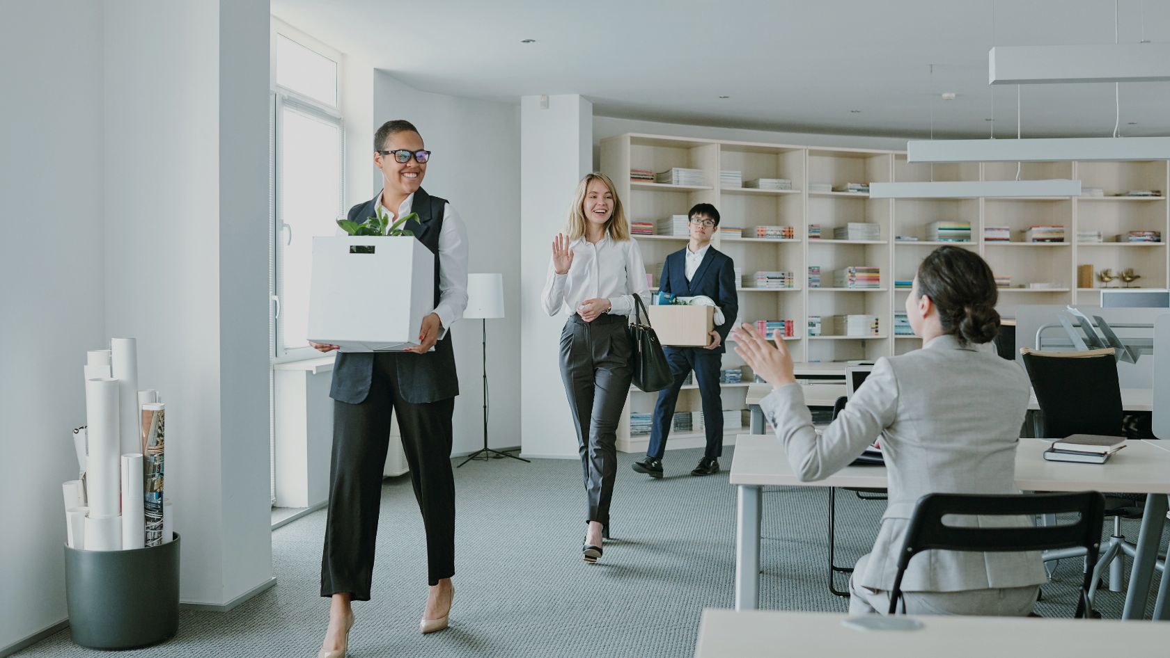 Three professionals walk through a bright office carrying moving boxes; the woman in front waves toward a seated colleague.