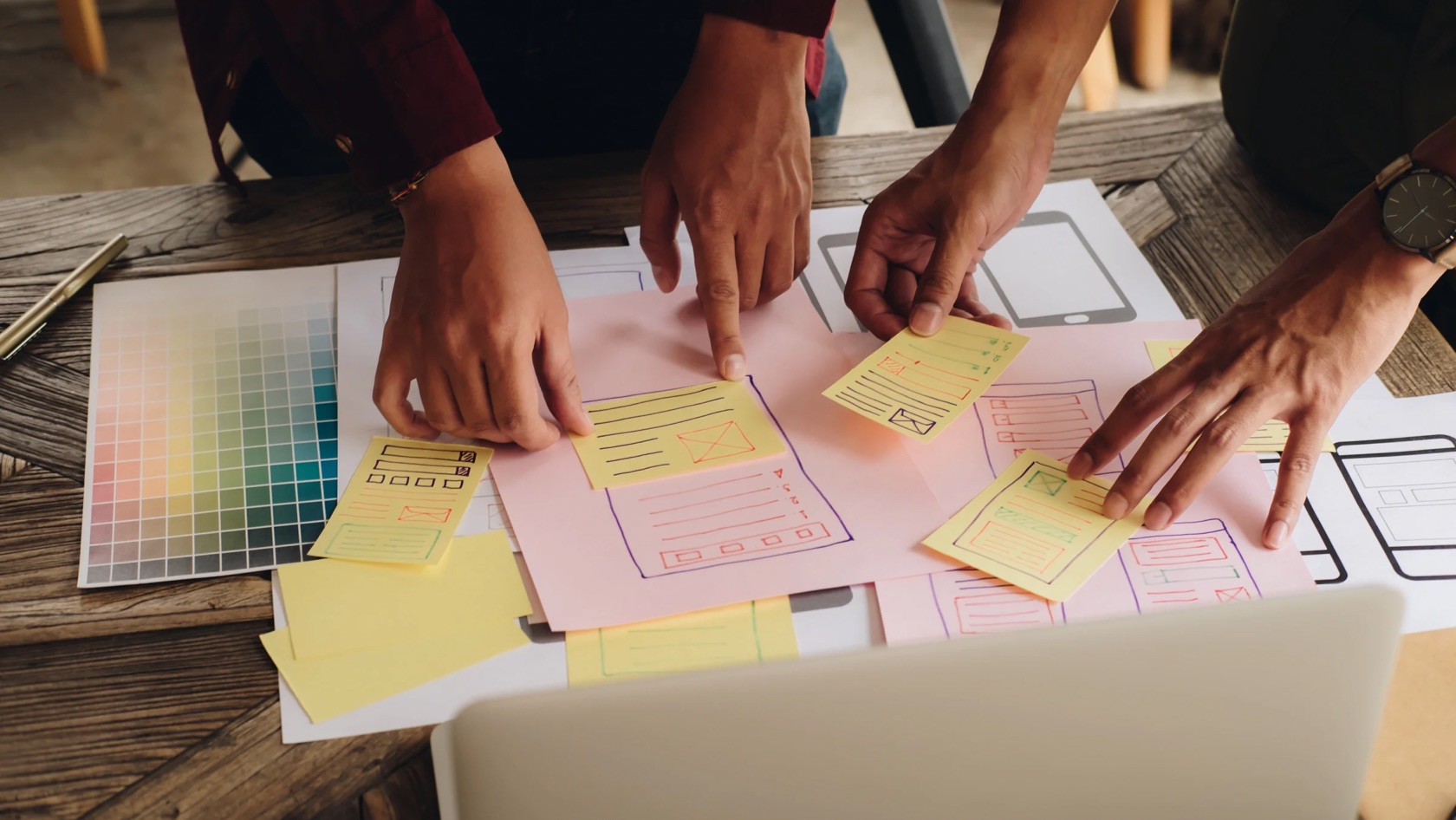 Hands arrange yellow sticky notes over app wireframe sketches on pink paper during a design session on a wooden table.