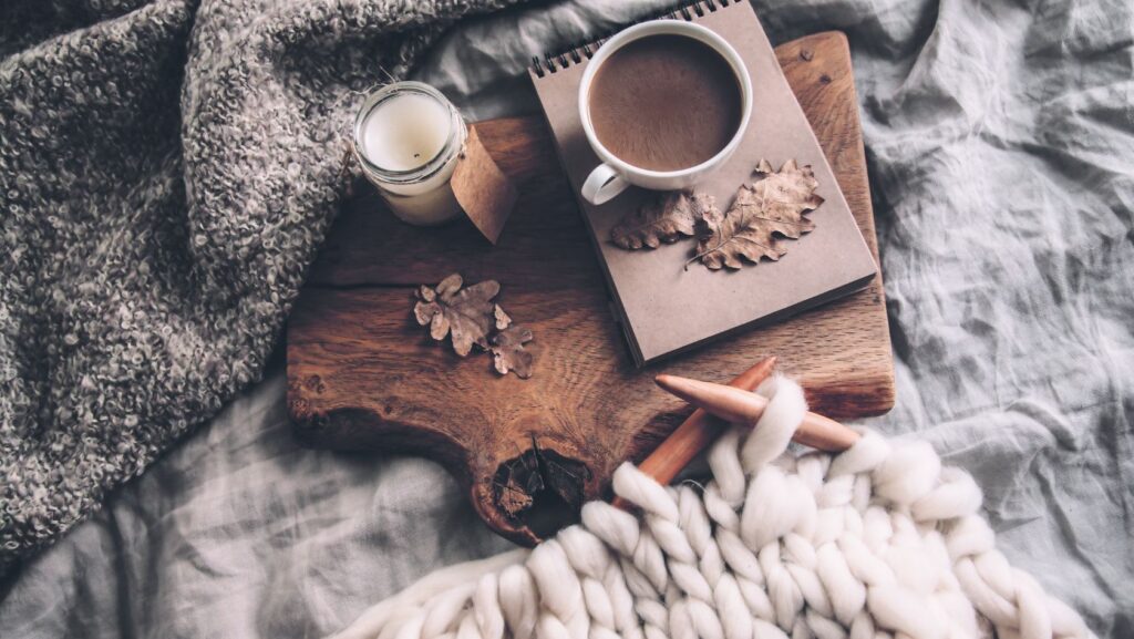 Cozy flat lay: mug of coffee, notebook with dried leaves, candle, and a wooden board topped with a chunky knit blanket and thick white yarn.