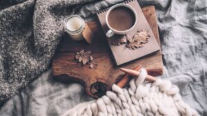 Cozy flat lay: mug of coffee, notebook with dried leaves, candle, and a wooden board topped with a chunky knit blanket and thick white yarn.