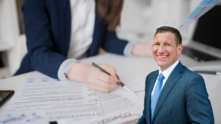 Smiling businessman in a blue suit sits at a desk with documents and a pen ready to sign the contract in front of him.