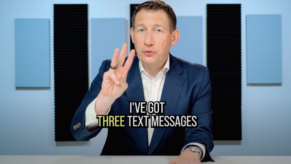 Man in a blue suit raises three fingers while saying 'I've got three text messages' in a studio setting with acoustic panels behind him.