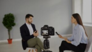 Camera on a tripod records an interview between a man and a woman in a bright room, both seated and blurred in the background.