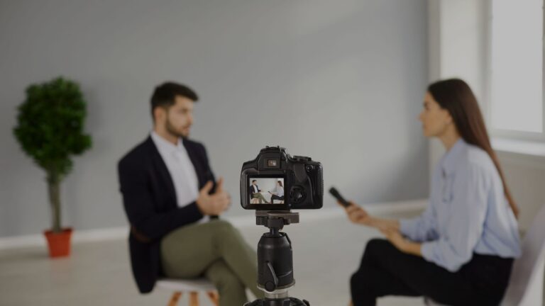 Camera on a tripod records an interview between a man and a woman in a bright room, both seated and blurred in the background.
