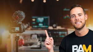 Smiling man in a black BAM shirt points at a DSLR on a tripod in a recording studio with a blurred workstation in the background.