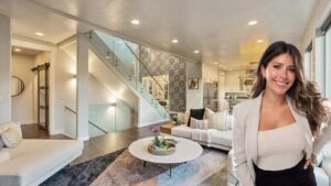 Smiling woman in a beige blazer stands in a bright open-concept living room with a glass staircase and white furniture.