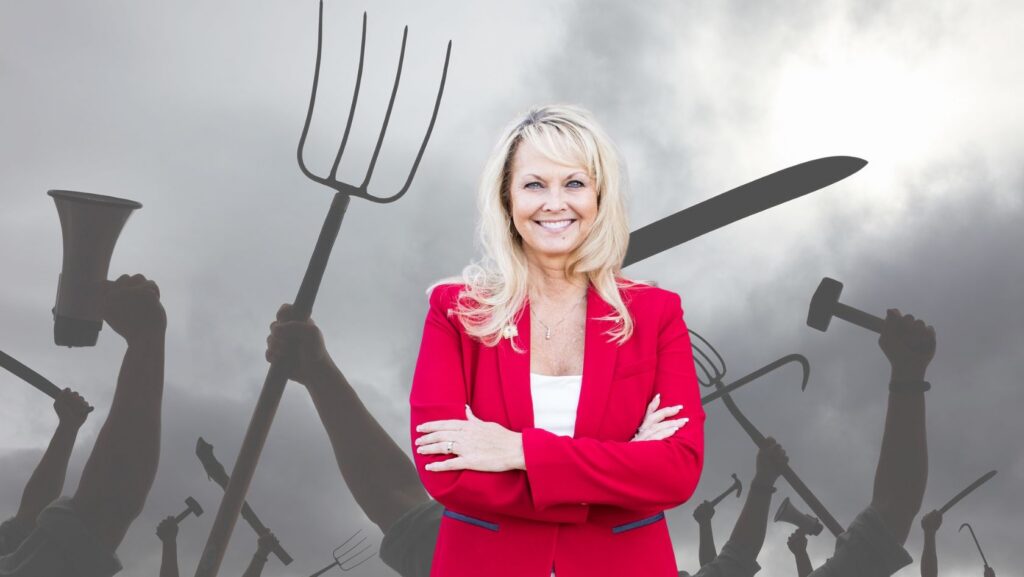 Smiling woman in a red blazer stands with arms crossed in front of a dark crowd holding various tools like pitchforks and hammers behind her, suggesting leadership at a protest or rally.