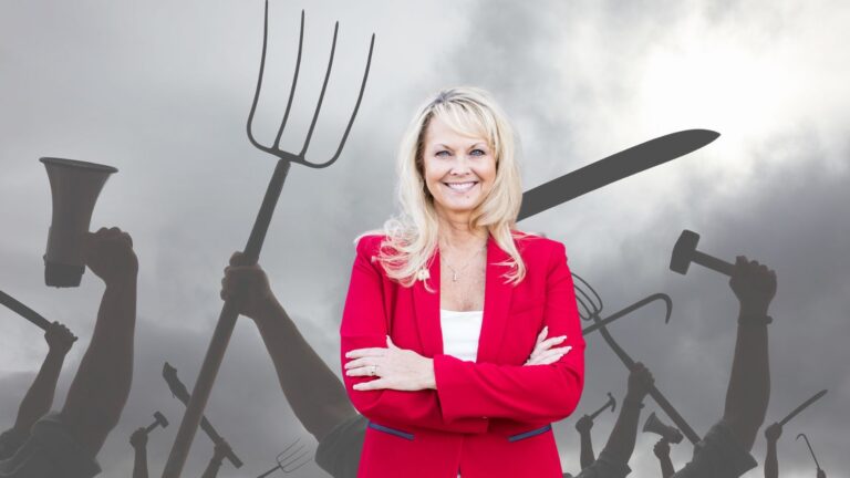 Smiling woman in a red blazer stands with arms crossed in front of a dark crowd holding various tools like pitchforks and hammers behind her, suggesting leadership at a protest or rally.