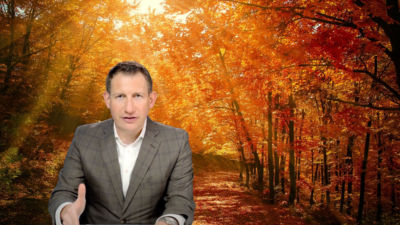 Man in a gray plaid suit sits at a desk or table, gesturing with his hand, with a sunlit autumn forest backdrop behind him.