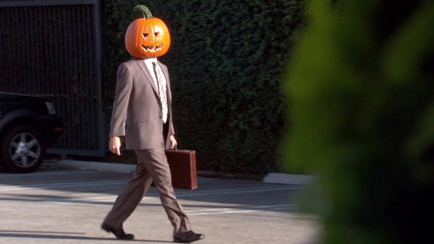 Person wearing a carved pumpkin as a head, in a brown suit and tie, carrying a briefcase outdoors in a parking area.