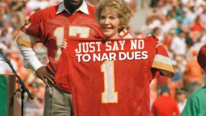 Smiling woman on a sports field holds a red football jersey with the text 'JUST SAY NO TO NAR DUES' and a crowd behind her, wearing red.