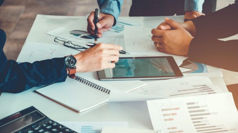 Two people collaborate over charts and documents on a table, with a tablet and notebook nearby.