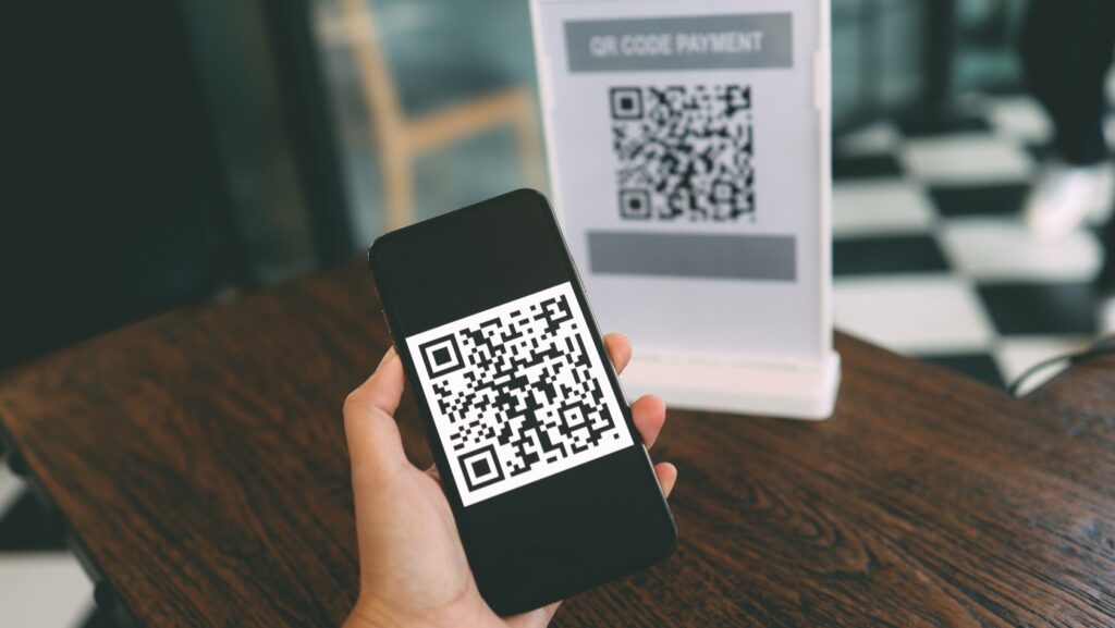 Hand holding a smartphone showing a QR code for payment, with a QR code payment sign visible in the background.