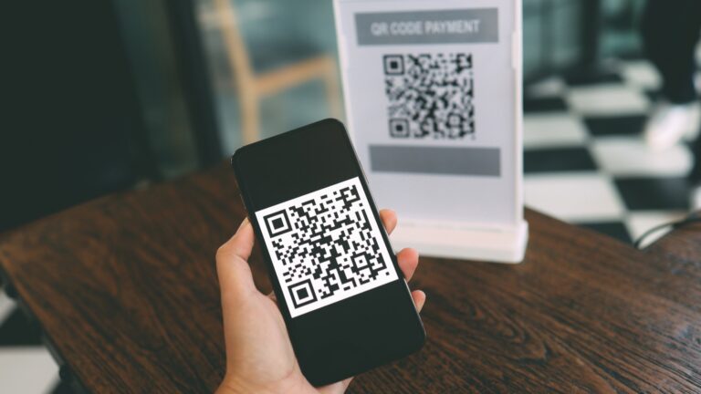 Hand holding a smartphone showing a QR code for payment, with a QR code payment sign visible in the background.