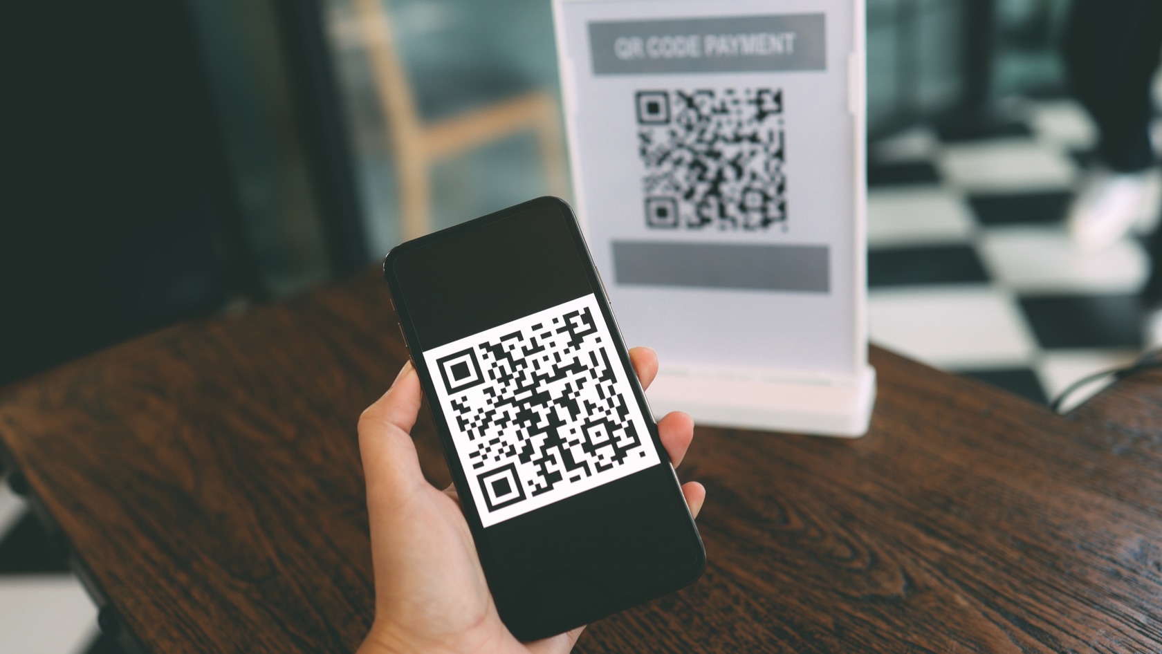Hand holding a smartphone showing a QR code for payment, with a QR code payment sign visible in the background.