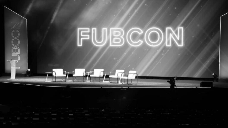 Conference stage with a podium on the left and six white chairs arranged for a panel, large neon 'FUBCON' backdrop behind.