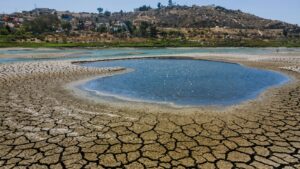 Cracked dry lakebed surrounding a blue circular pool, with a hillside town in the distance under a clear sky.