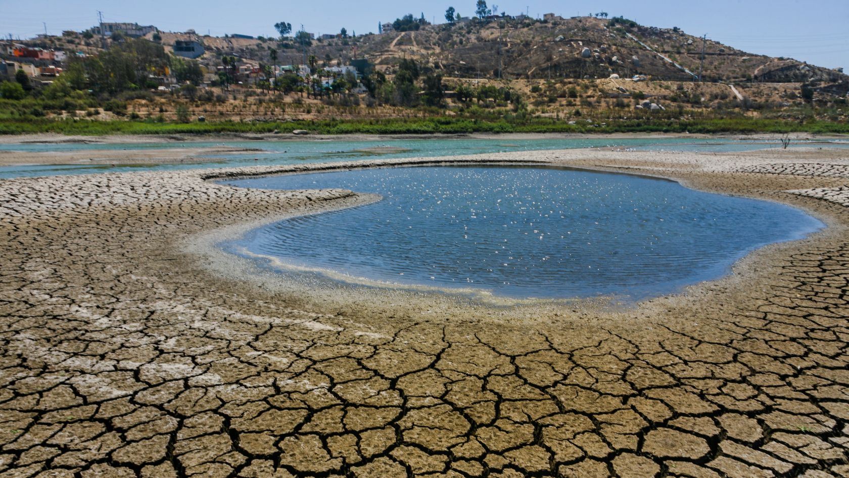 Cracked dry lakebed surrounding a blue circular pool, with a hillside town in the distance under a clear sky.