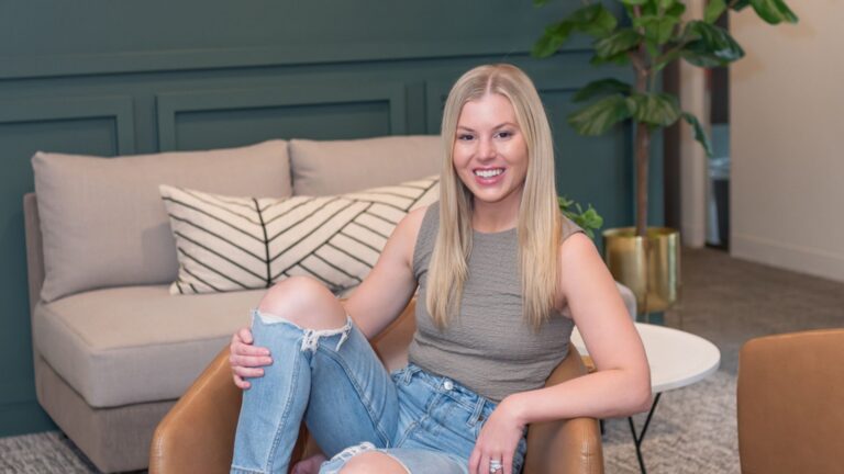 Smiling woman with long blonde hair sitting in a leather chair in a modern living room gardened by a teal wall behind her.
